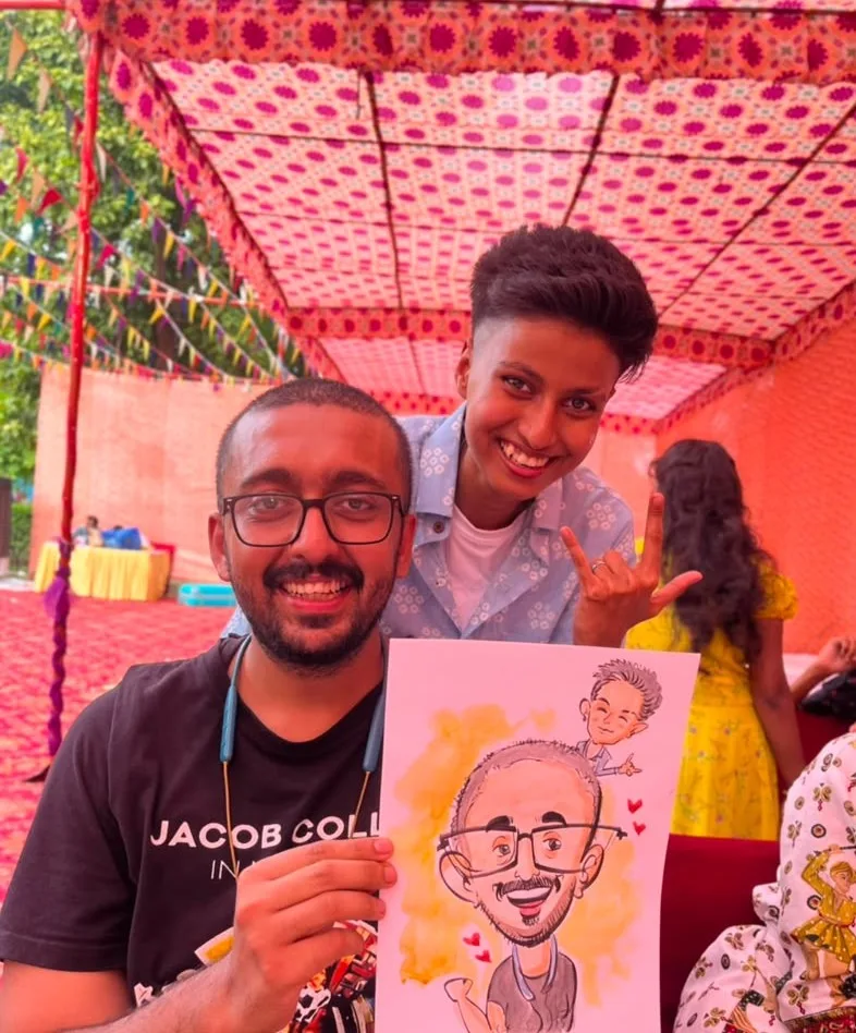 A man and a woman holding up the caricature just drawn of the two of them, at an outdoor function under a pink patterned canopy