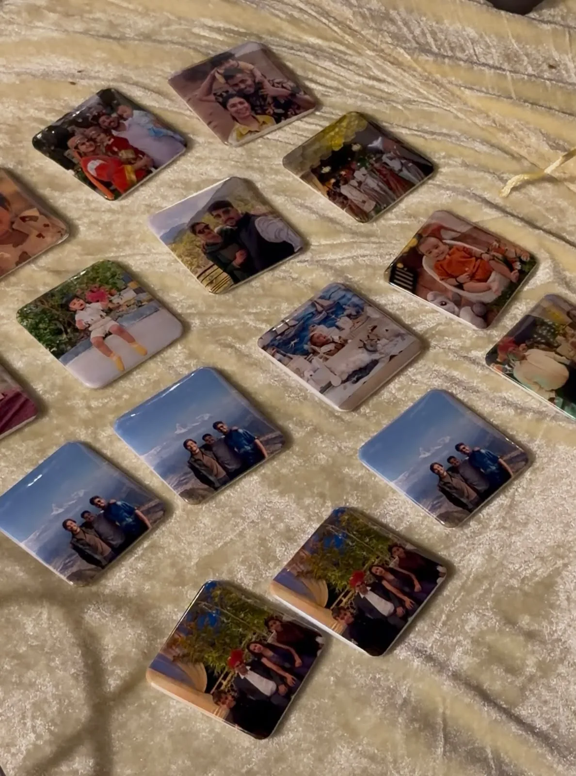 Glossy square photo magnets with rounded corners laid out on a cream cloth, printed with family photos, a baby portrait and a group at a mountain viewpoint
